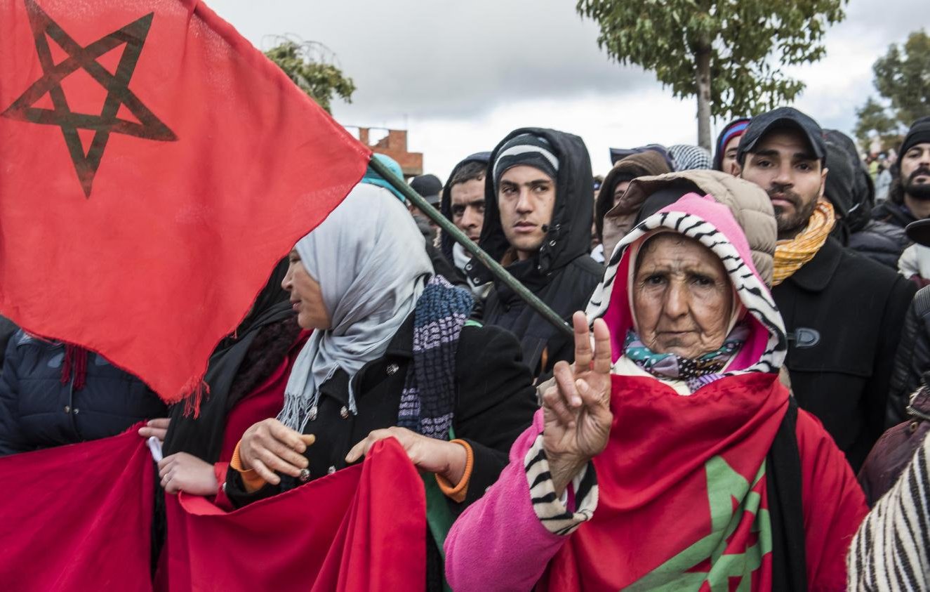 The image depicts a group of people participating in a demonstration. In the foreground, an older woman holds a flag featuring the Moroccan national emblem and makes a peace sign with her hand. She is surrounded by others who are also holding flags and banners, all dressed for colder weather. The atmosphere suggests a sense of solidarity among the demonstrators, with the crowd positioned against a backdrop of cloudy skies.