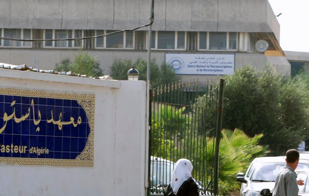 The image depicts a building with signage indicating it is the "Institut Pasteur" in Algeria. In the foreground, there is a decorative wall with Arabic text, likely the name of the institution, along with some greenery. You can also see several people walking along a path near the entrance. The architectural style of the building appears modern and utilitarian.