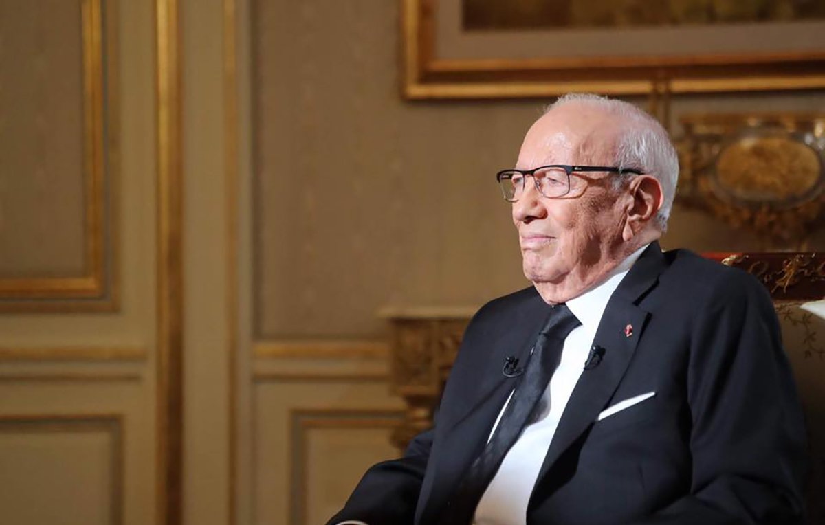 The image depicts an elderly man sitting in a formal setting. He is wearing glasses and a black suit, along with a black tie. The background features tasteful decor with intricate designs and warm colors, suggesting an elegant environment. The man appears to be engaged in thought or reflecting, with a calm and serene expression on his face.