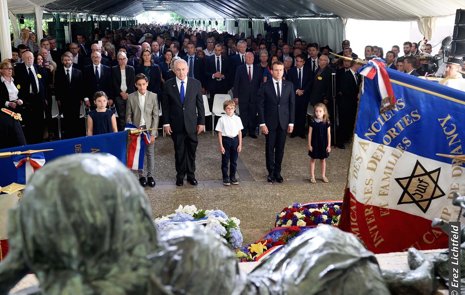 The image depicts a solemn gathering under a tent, likely at a memorial or commemorative event. In the foreground, there are several children and adults standing attentively. Prominent figures, possibly including political leaders, are present, and flags, including one with a star emblem, are displayed around them. The atmosphere appears respectful and reflective, suggesting it is a tribute to those who have passed or a celebration of their memory. Floral arrangements are visible, adding to the somber yet respectful tone of the scene.