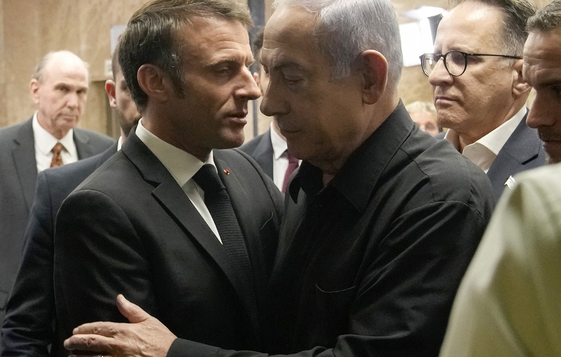 The image depicts two men in close proximity, engaged in a serious conversation. One man is dressed in a formal suit and tie, while the other is wearing a black shirt. They appear to be in an indoor setting, surrounded by several other individuals in the background, who are also dressed in formal attire. The atmosphere looks tense or important, suggesting a significant discussion is taking place.