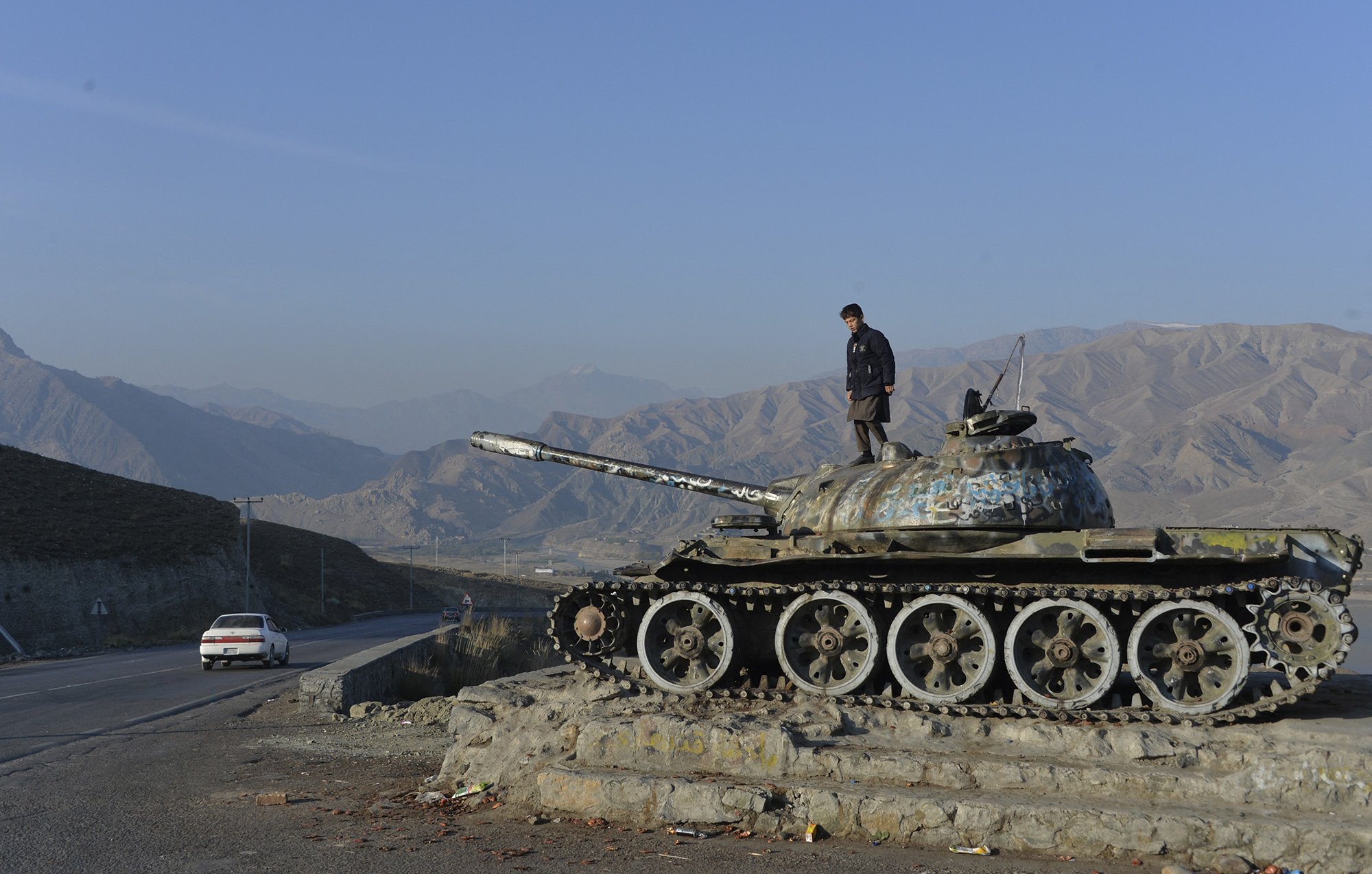 The image depicts a person standing atop an old tank, which is positioned on a small rise alongside a road. The tank appears weathered and has a layer of rust. In the background, there are rolling mountains under a clear blue sky, creating a stark contrast with the tank's dilapidated condition. A car can be seen on the road nearby, suggesting a sense of movement in this otherwise desolate landscape. Overall, the scene conveys themes of history and the passage of time in a rugged environment.