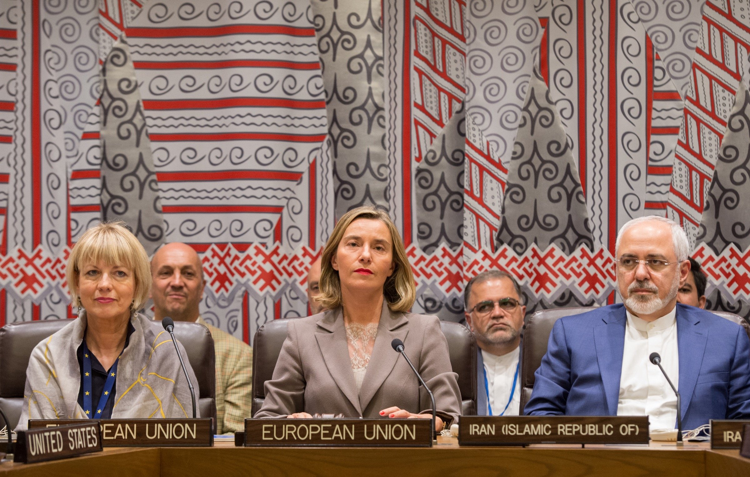 The image shows a formal setting, likely a meeting or conference related to international diplomacy. In the foreground, there are three individuals seated at a table, each identified by nameplates indicating their affiliations: the United States, the European Union, and Iran. The woman in the center, dressed in a professional outfit, appears to be the main speaker or moderator. Behind them, several other individuals are visible, suggesting a diverse group of participants in the meeting. The backdrop features a decorative pattern, adding a cultural context to the setting.