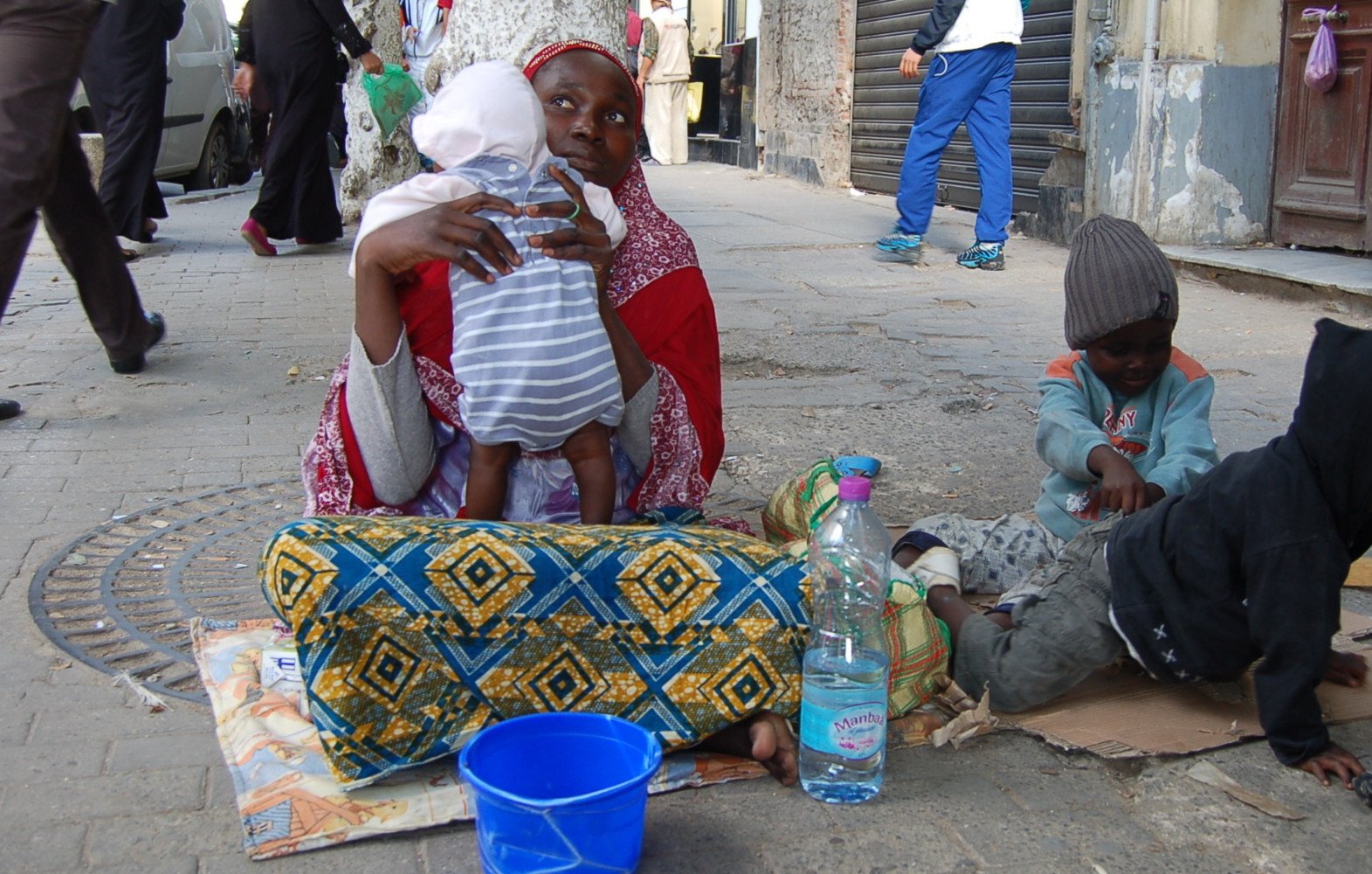 The image depicts a scene on a city street where a woman is seated on the ground with a baby in her arms. She appears to be wearing traditional clothing, characterized by vibrant colors and patterns. Nearby, two young children are playing on the pavement; one is sitting in a casual posture, while the other is occupied with an object. There is a blue bucket beside them and a plastic bottle of water. The background includes other people walking by and buildings that suggest an urban environment.