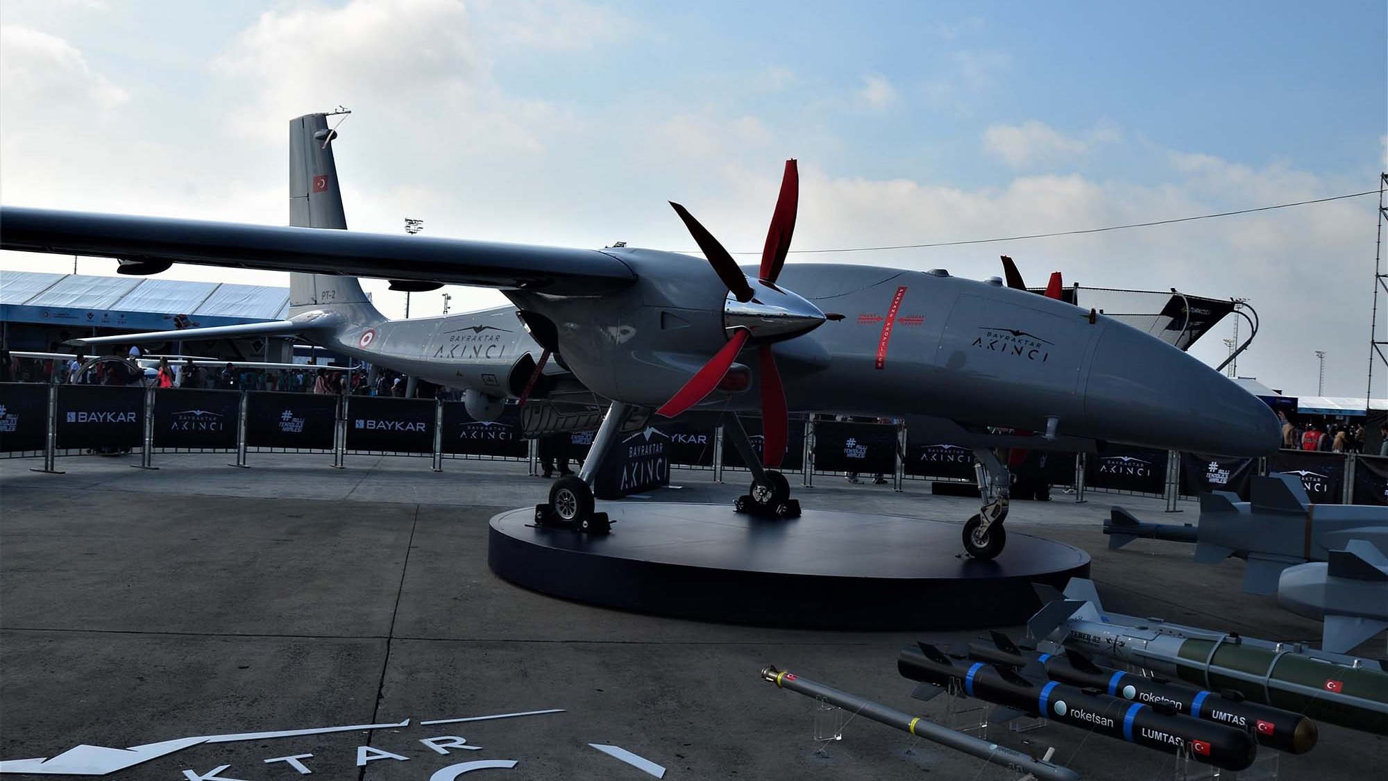 The image features a military drone displayed prominently on a platform. The drone has a sleek, aerodynamic design with multiple red propellers. It is painted in a gray color, which is common for military equipment. In the foreground, there are several missiles and munitions arranged neatly, showcasing the drone's armament potential. The background includes a tent structure and additional drone models, suggesting this is part of an exhibition or airshow. The branding on the drone indicates it is a product of Baykar, referencing a specific model or variant likely designated for military use.