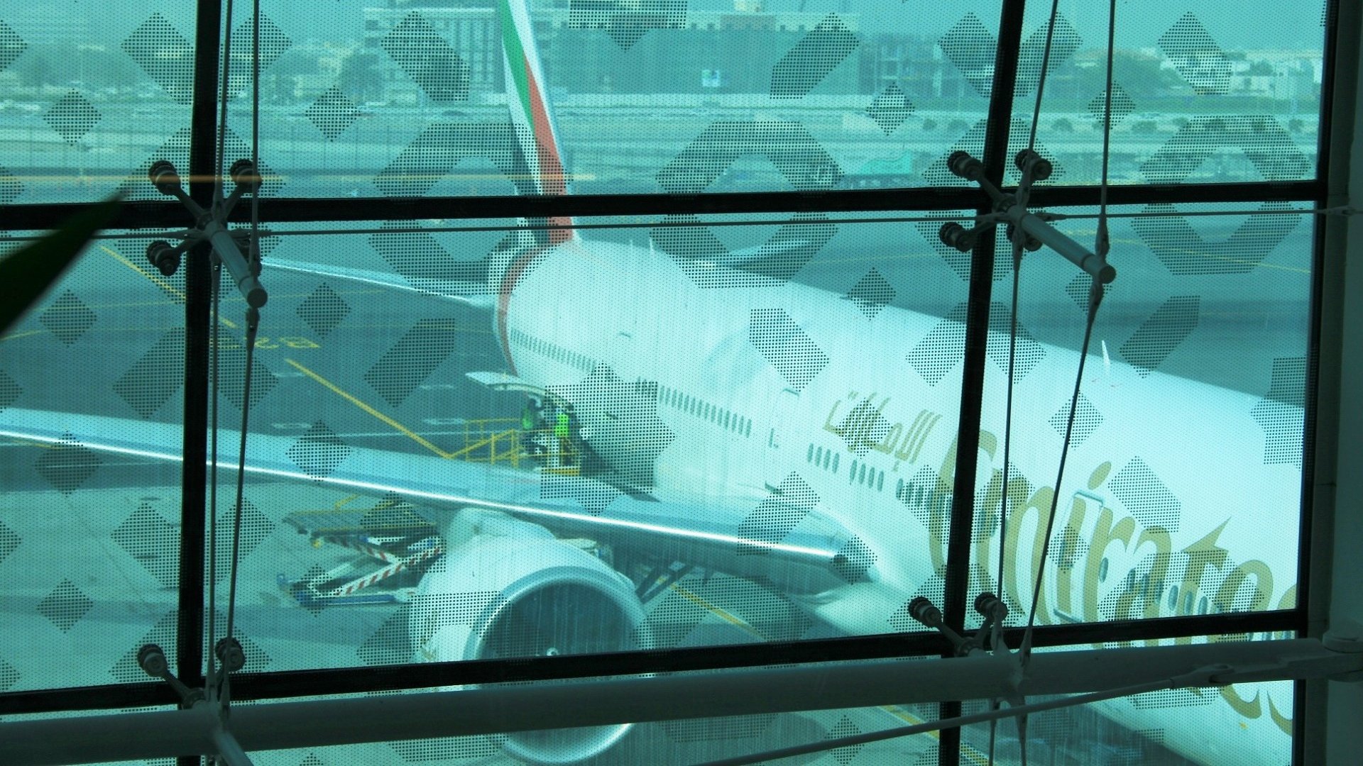 The image shows an airplane belonging to Emirates Airlines parked at an airport. The aircraft appears to be a large model, likely a Boeing 777, as indicated by the shape of the fuselage and engines. You can see maintenance personnel working on the plane, and the background features airport buildings and infrastructure. The scene is framed by a glass wall, which has a patterned overlay, adding an artistic element to the view. The overall atmosphere seems busy and operational, typical for a major airport.