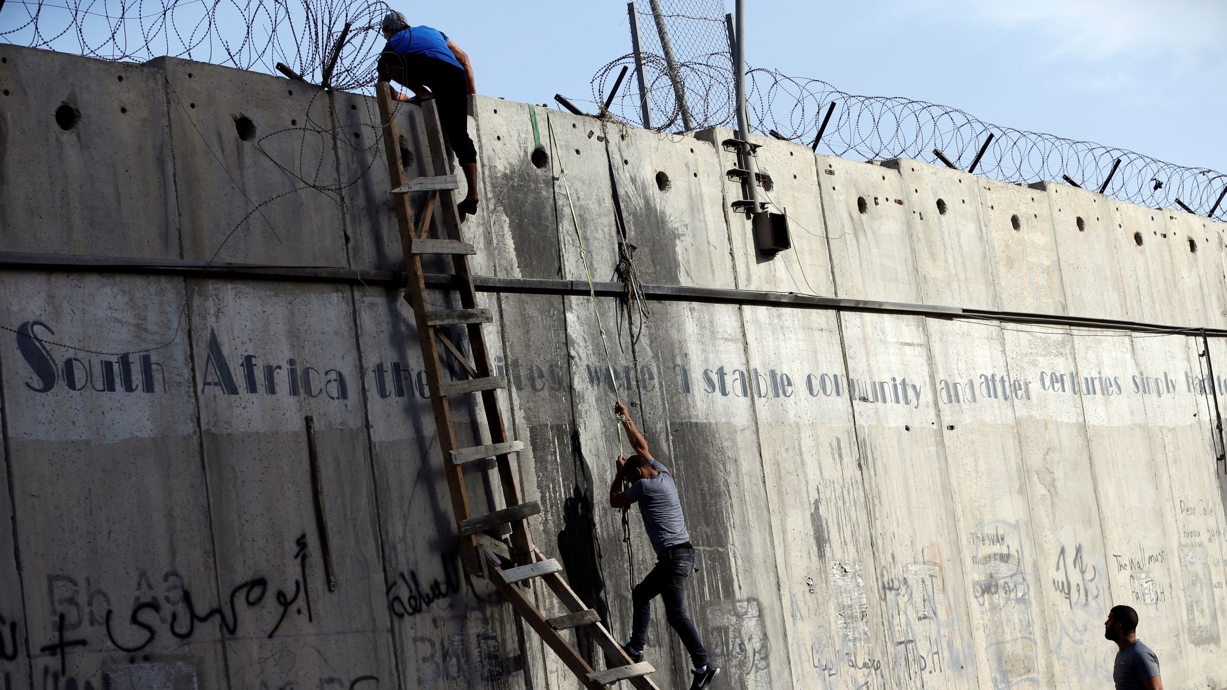 The image shows two individuals climbing a tall concrete wall, which features barbed wire at the top. A ladder is positioned against the wall, aiding their ascent. The wall bears graffiti, including text in multiple languages, and there is a phrase that mentions "South Africa" likely in the context of stability and historical struggles. The setting appears tense, capturing a moment of movement against a backdrop of urban infrastructure.