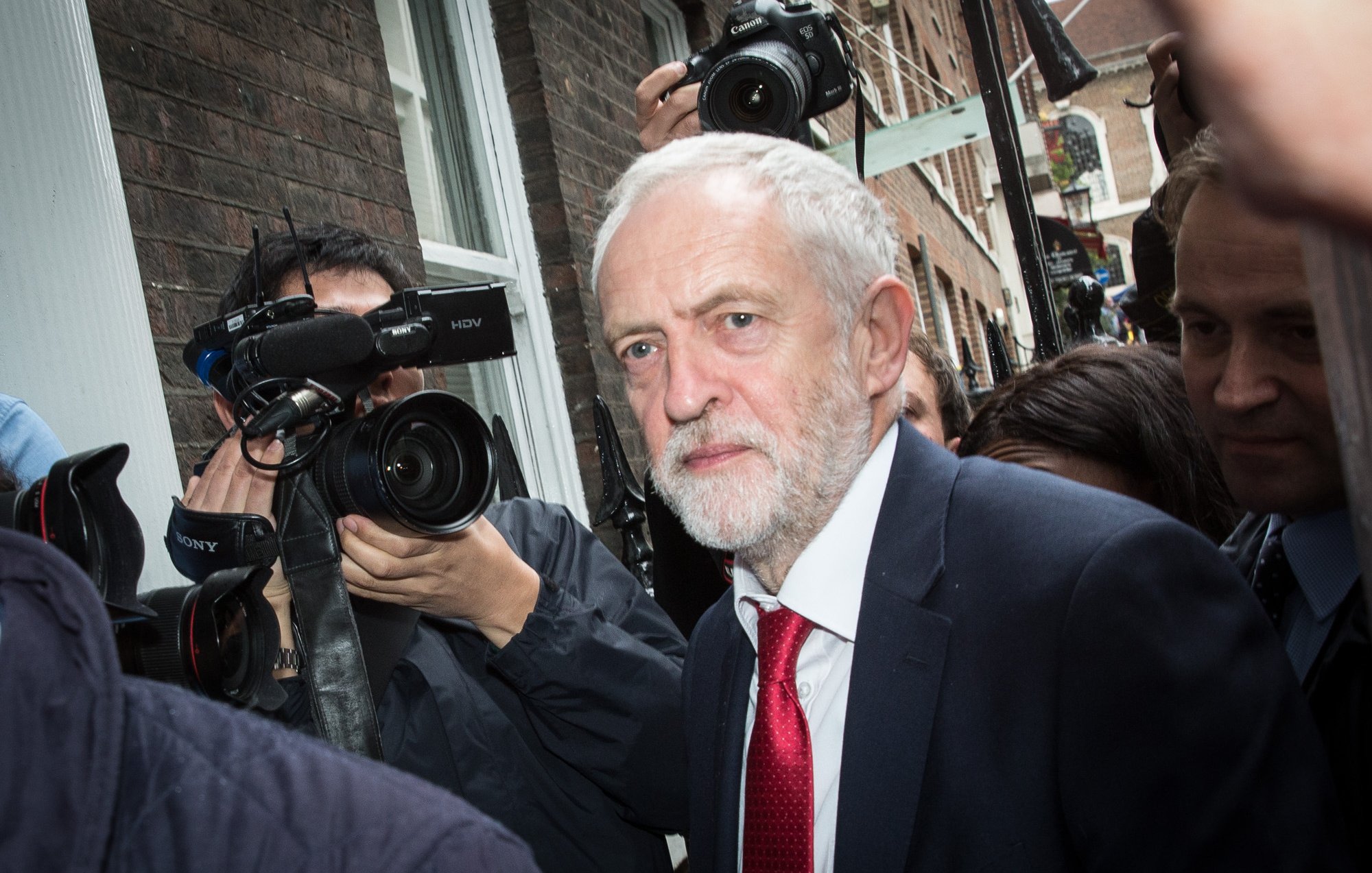 The image shows a man with gray hair and a beard, dressed in a dark suit and a red tie, walking through a crowd. He appears to be in a busy outdoor setting, surrounded by photographers and cameras capturing the moment. The buildings in the background suggest an urban environment. The overall atmosphere seems to convey a sense of public attention and media interest.