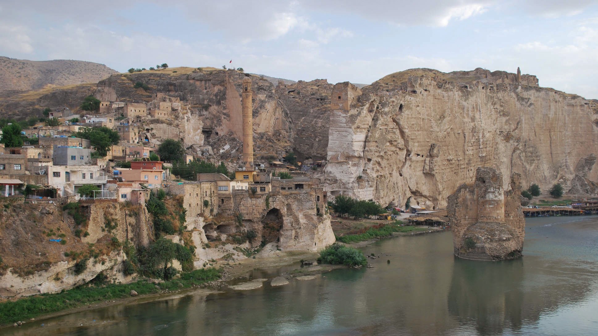 The image depicts a scenic landscape featuring a river flowing through a valley. On the left side, there are rocky cliffs and remnants of ancient structures, hinting at a historical settlement. The area appears to be a mix of natural and man-made elements, with a few buildings clinging to the hillside. In the background, there are more cliffs and a prominent tower-like structure, suggesting a rich history and possibly a fortified site. The sky is partly cloudy, adding to the serene atmosphere of the scene.