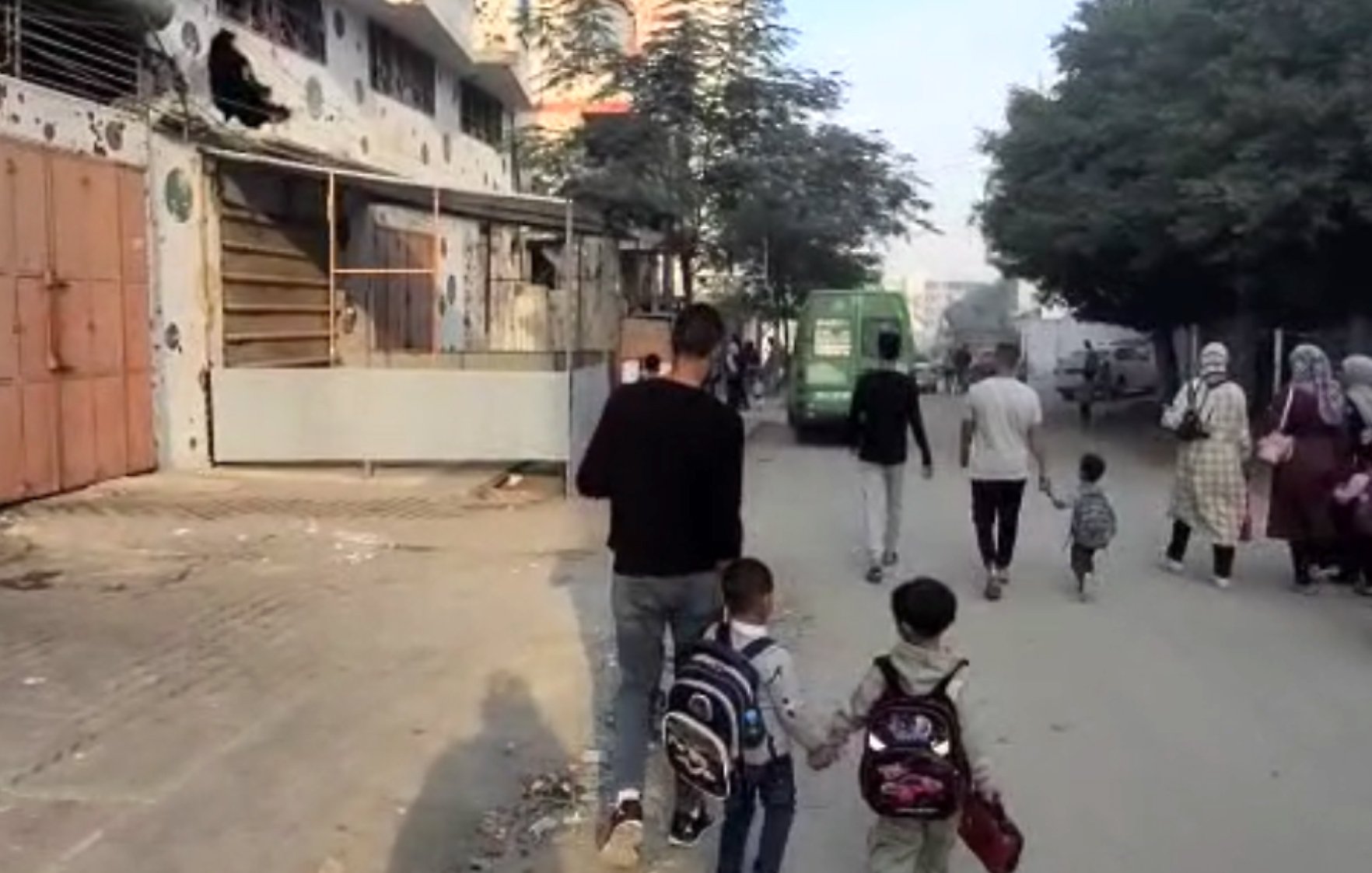 A man walks with two children on a dusty street, while others stroll nearby. Buildings line the path.