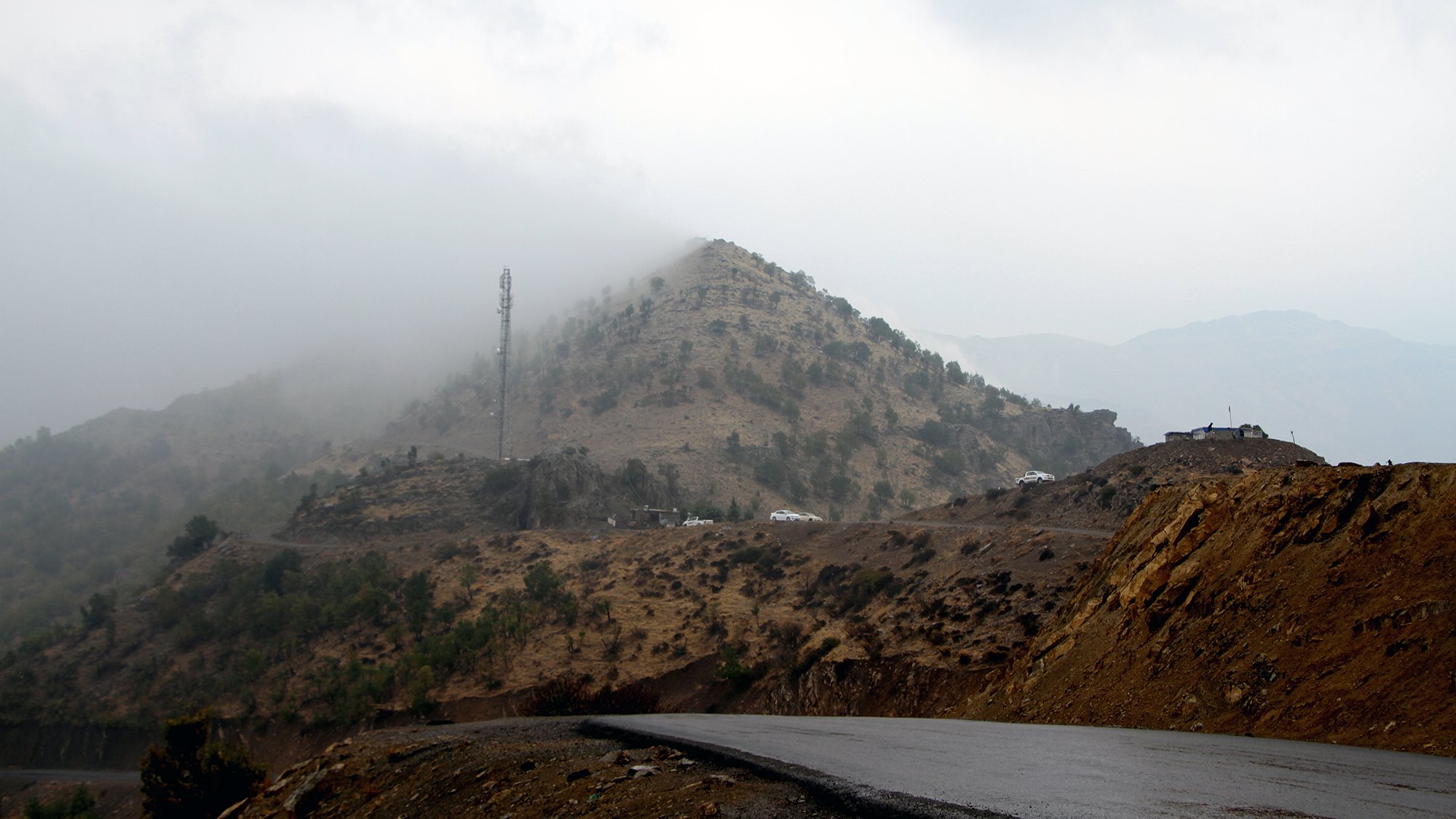 The image depicts a winding road leading through a mountainous landscape. In the background, a hill covered with low vegetation is partially obscured by mist or fog, creating a mysterious atmosphere. Atop the hill, there is a communication tower, and some vehicles are visible near the summit. The foreground features a dark, asphalt road that curves, suggesting a remote and rugged terrain. The overall scene conveys a sense of tranquility and solitude in nature.