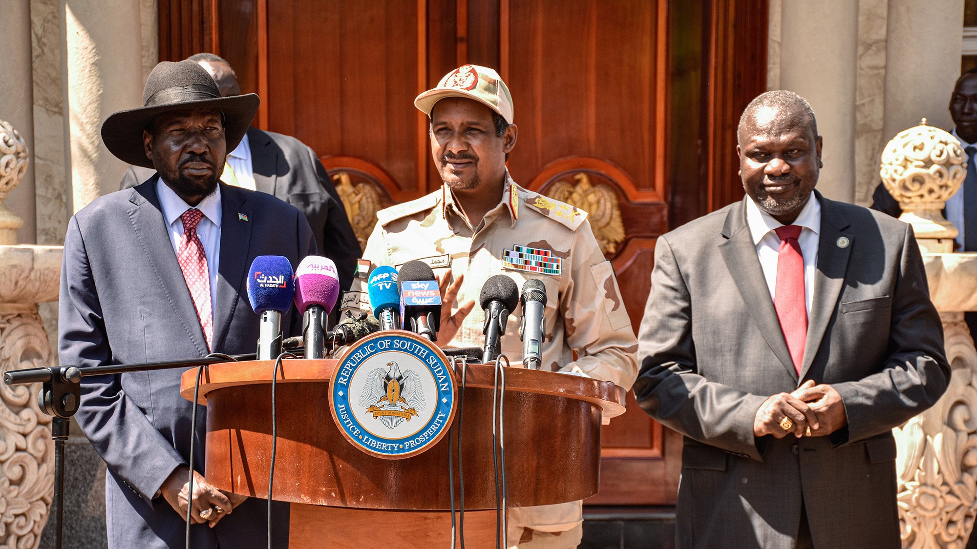 The image shows a group of three men standing behind a wooden podium. The man on the left is wearing a black hat and a suit with a red tie. The man in the center is wearing a military uniform with various insignia and a cap. The man on the right is dressed in a dark suit. They appear to be at an official event, likely addressing the media, with microphones positioned in front of them and a decorative building in the background.