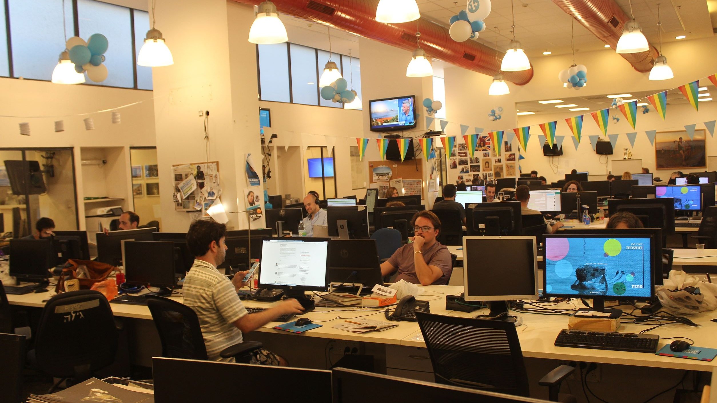 The image shows an open office space with several workstations. There are multiple desks equipped with computers, where people are engaged in various tasks. The decor includes colorful balloons and banners hanging from the ceiling, suggesting a festive atmosphere. Large windows allow for ample natural light, and the overall ambiance seems lively and collaborative, with employees working both individually and in small groups.