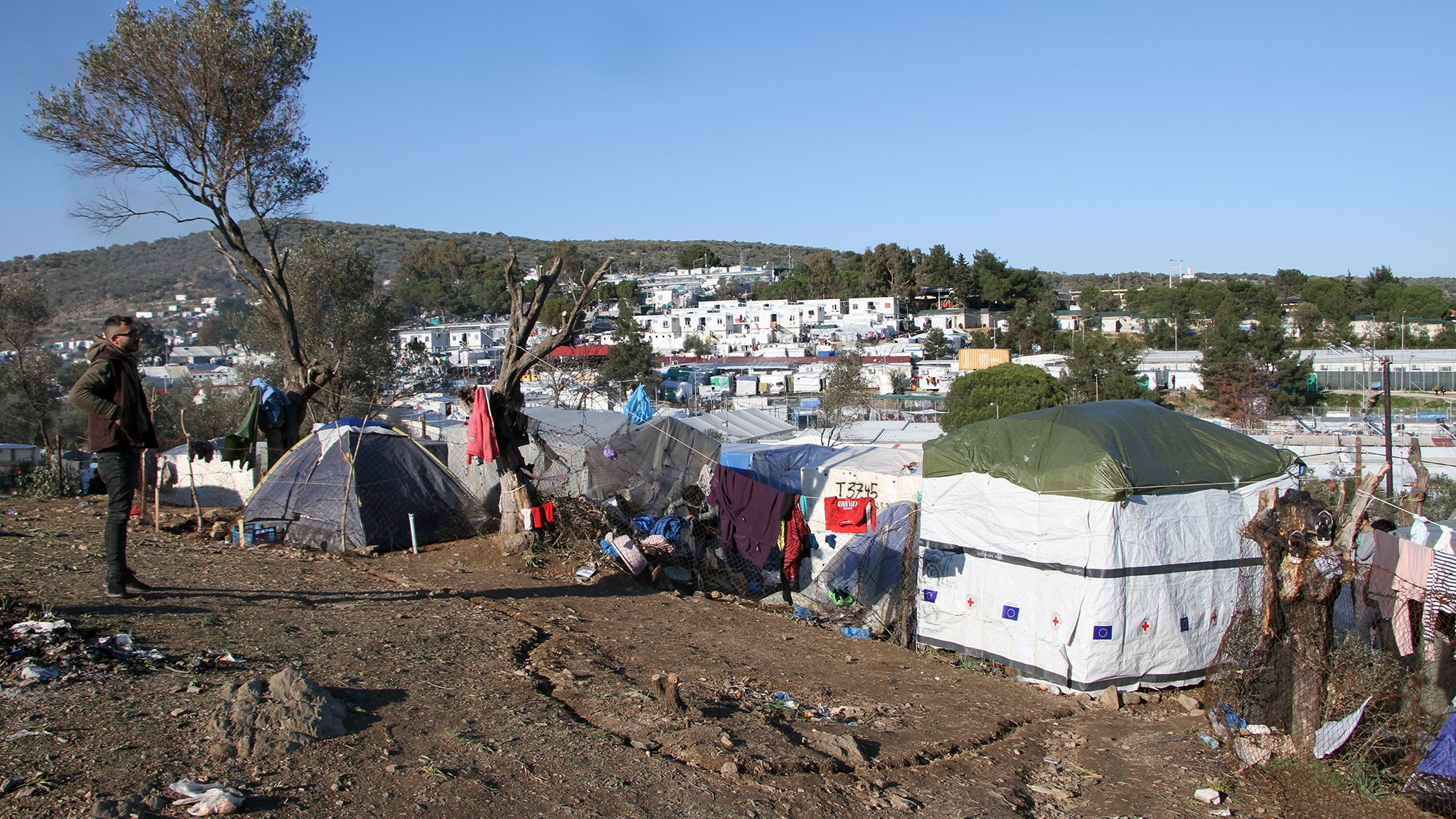The image depicts a makeshift settlement in a rural or semi-urban area. Numerous tents are arranged on a slope, suggesting a temporary living situation. The landscape features sparse vegetation and a few trees, while in the background, there are more established buildings, possibly residences or facilities. The scene conveys a sense of hardship, with signs of disarray and poverty surrounding the tents. The sky is clear with little cloud cover, indicating a sunny day. A person stands in the foreground, observing the surroundings.
