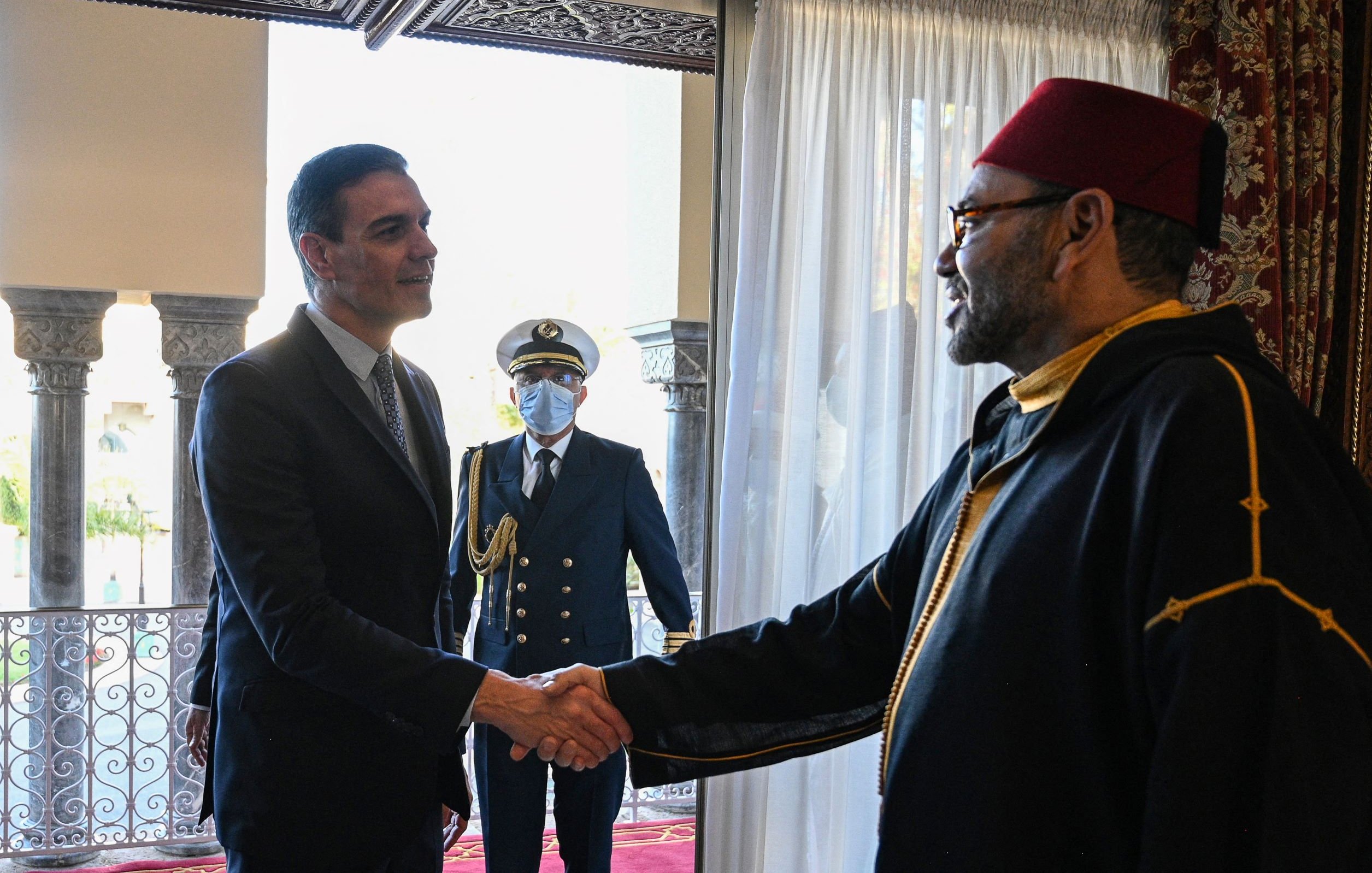The image depicts a meeting between two men. One man, dressed in a suit, is shaking hands with the other man, who is wearing traditional attire, including a red fez. In the background, a military figure stands in uniform, observing the handshake. The setting appears to be an elegantly decorated room with large windows and elaborate architectural details.