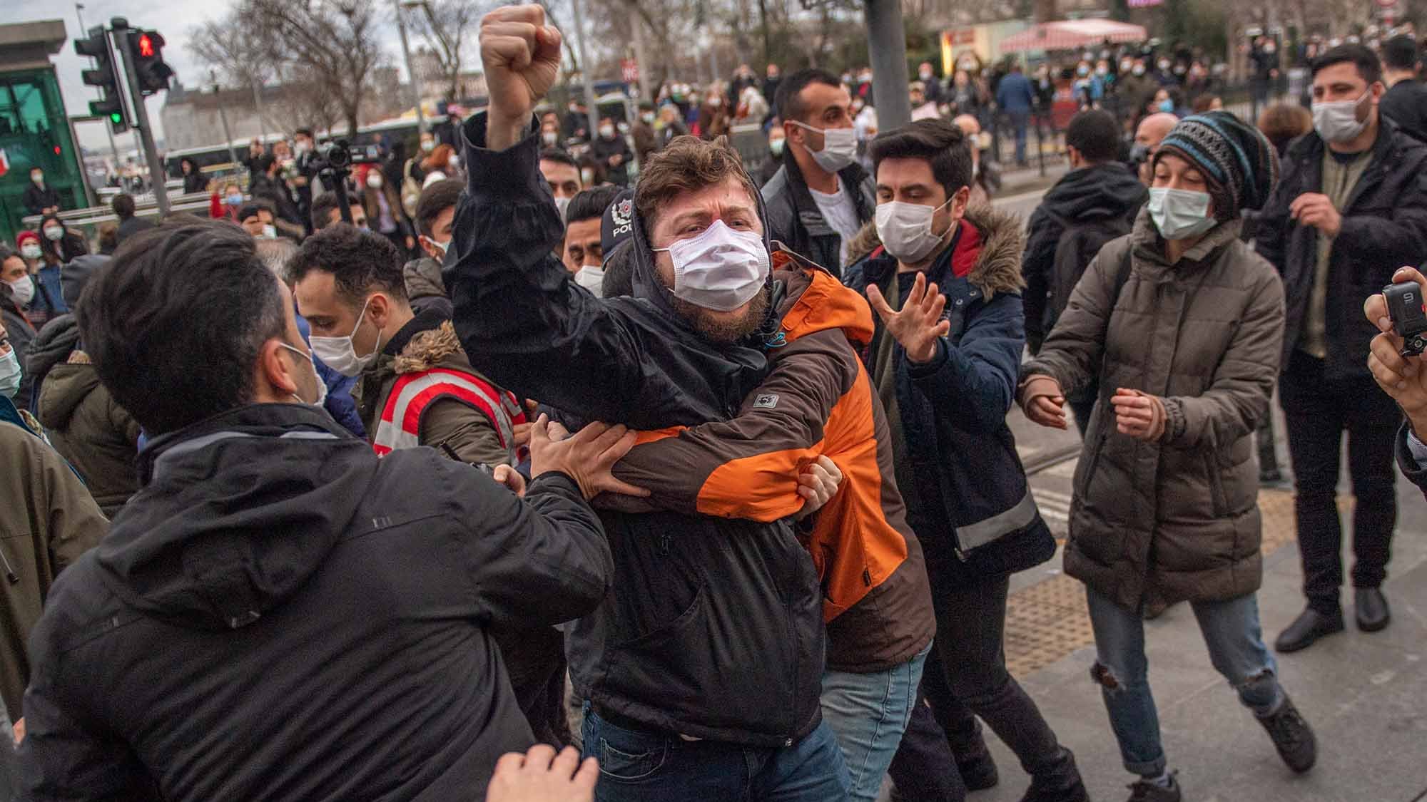 The image depicts a scene of confrontation during a protest or demonstration. Several individuals are gathered, with some appearing to engage in a physical altercation. One person, wearing a black jacket and a mask, is raising a fist, indicating a strong emotional response. People around them seem to be trying to intervene or separate those involved in the conflict. The setting appears to be an urban area, likely near a traffic light, with a backdrop of buildings and other demonstrators. The atmosphere seems tense and charged.