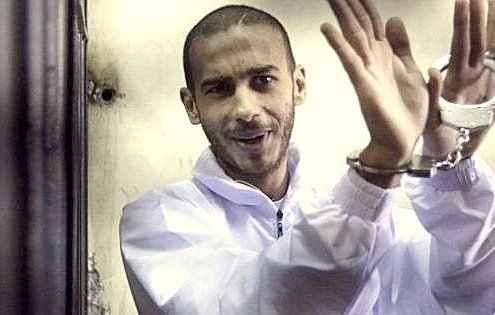 The image depicts a man in a white jumpsuit, appearing to smile or show a light-hearted expression. He is wearing handcuffs and seems to be gesturing with his hands, possibly in a playful or confident manner. The background appears to be a dimly lit space, which adds to the atmosphere of the scene.