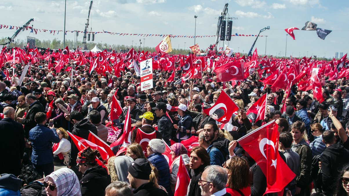 The image shows a large crowd gathered in an outdoor setting, with many people holding Turkish flags. The atmosphere appears to be lively and energetic, with individuals visibly engaged in the event. Some are holding banners, while others are raising their flags high. The background features clear skies and some structures, possibly related to the event or gathering. Overall, it depicts a significant public demonstration or celebration, likely related to a national or cultural event in Turkey.