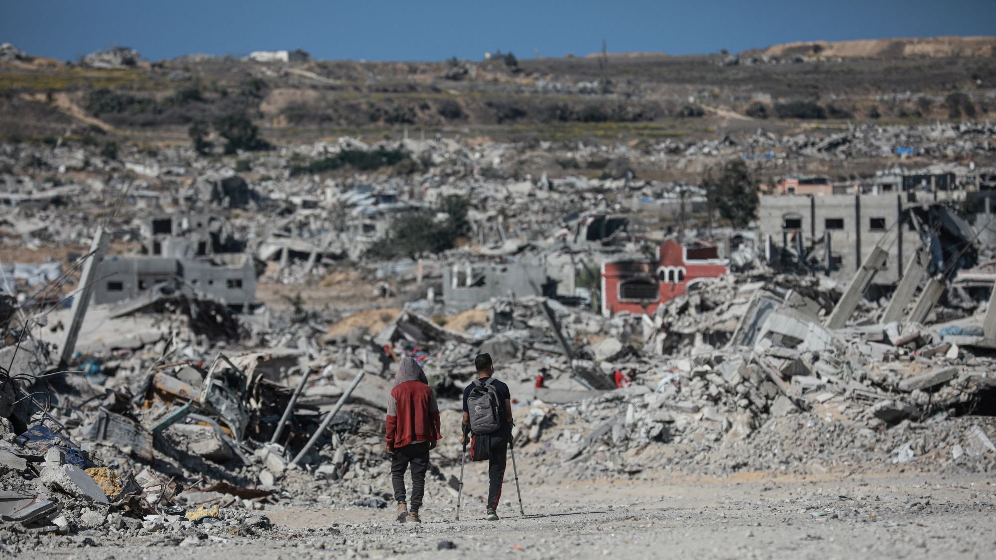 Two people walk through a vast landscape of destruction and rubble, with collapsed buildings around them.