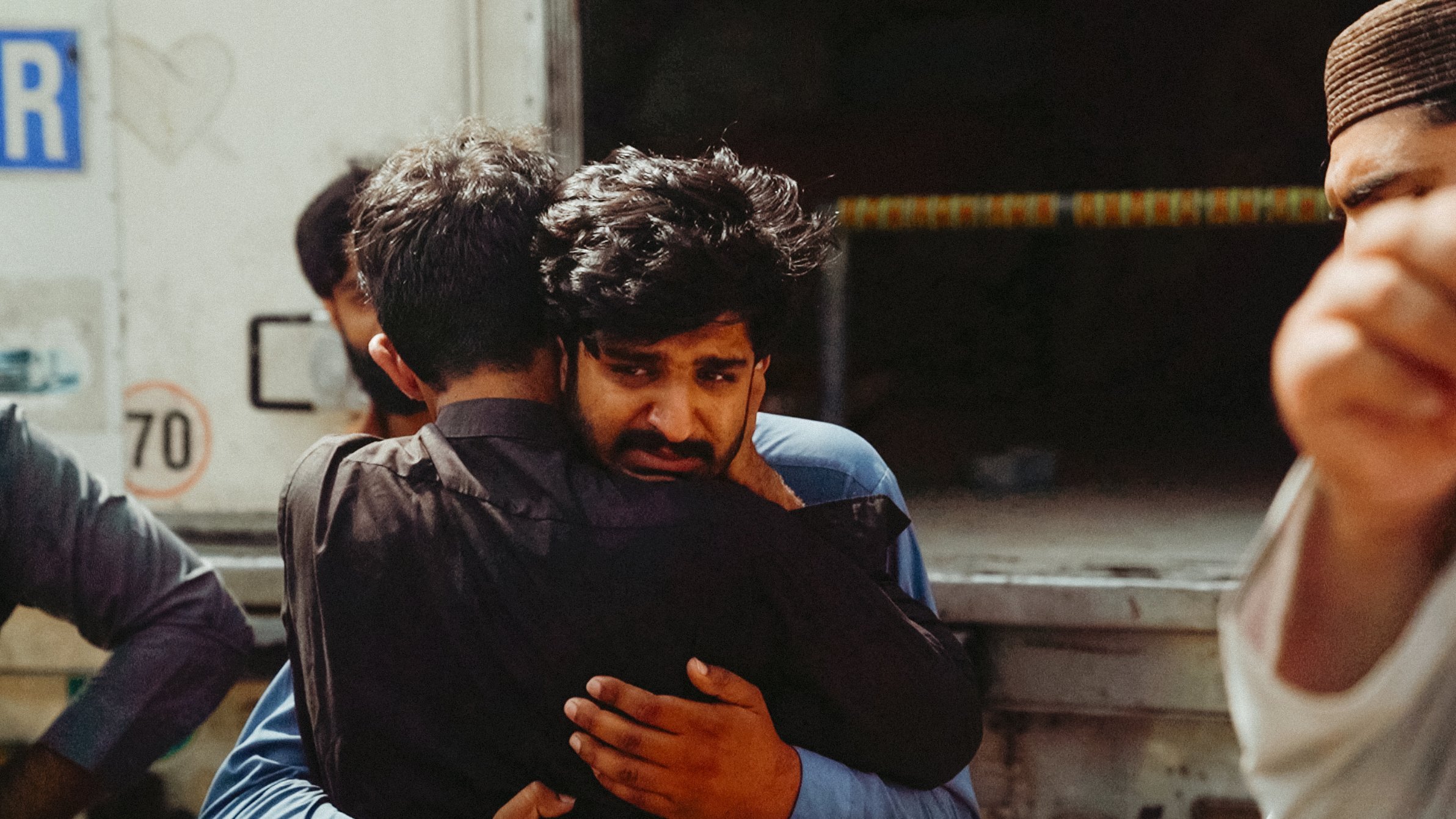 Two men embrace tightly, one looks visibly emotional, surrounded by others in a somber setting.