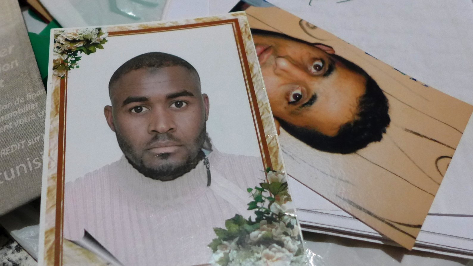 The image shows two photographs. One is a formal portrait of a man with short hair, wearing a light-colored sweater. The background contains decorative elements like flowers. The second photo appears to be a more candid or casual shot of another man, who has dark, curly hair and a neutral expression. Both photos are placed together on a surface that includes some papers and miscellaneous items.
