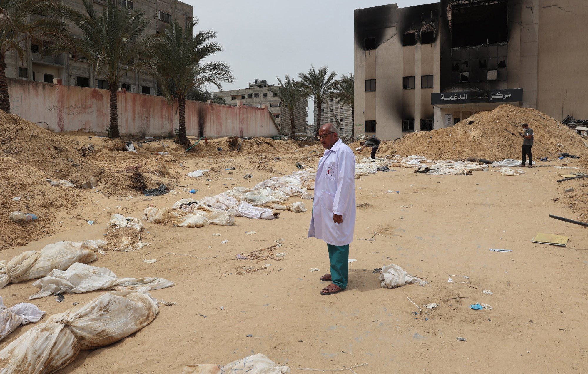 The image depicts a scene in a sandy area where several bodies, covered in white shrouds, are lying on the ground. In the foreground, a man wearing a white coat stands solemnly. In the background, there are damaged buildings, some of which appear to be burned. Palm trees line the area, and the overall atmosphere seems somber and grim, indicating a setting of conflict or disaster.