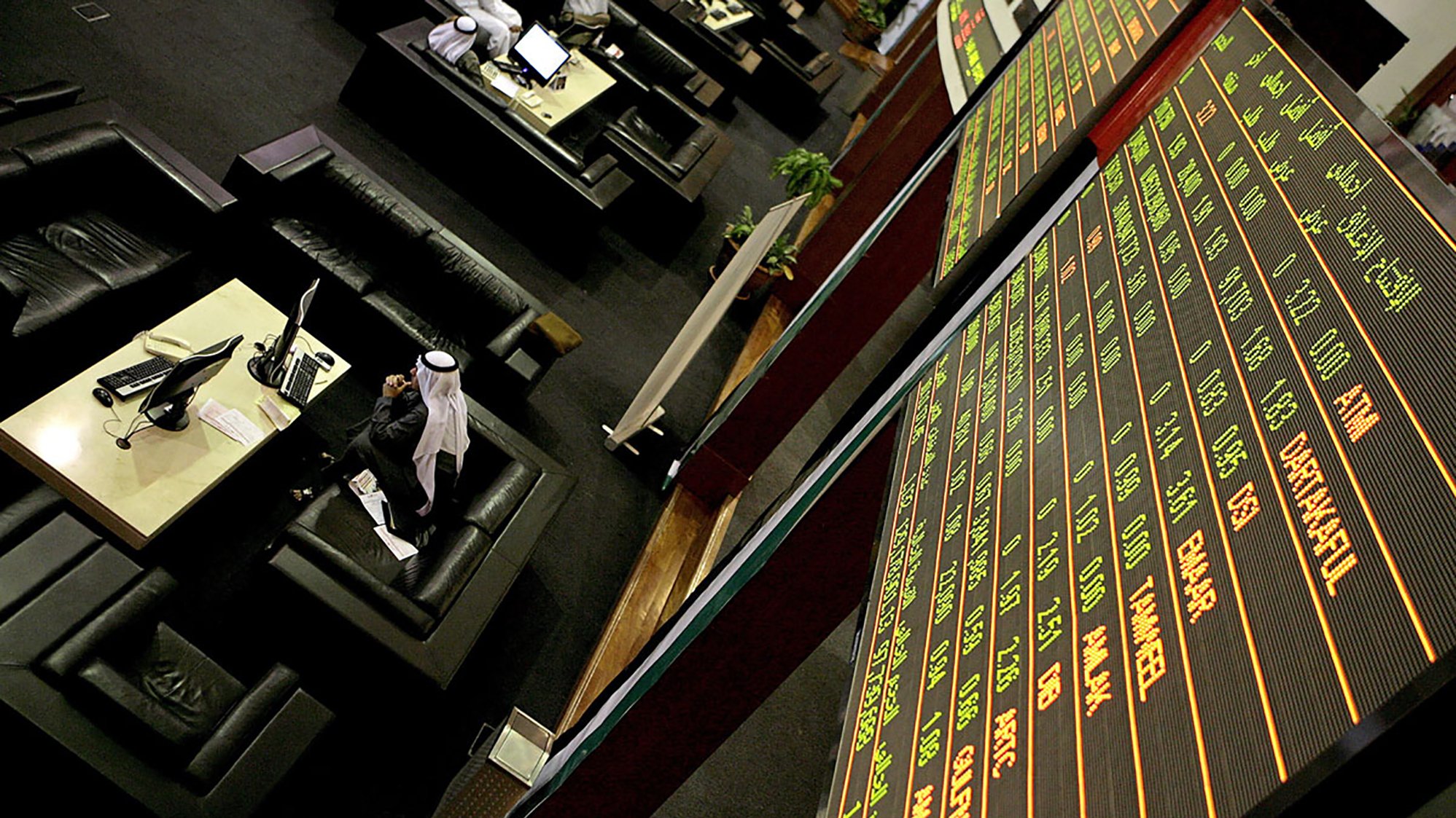 The image depicts a financial trading environment, likely within a stock exchange. In the foreground, there is a large electronic display board showing various financial data, such as stock prices and indices, with green and red text indicating changes in value. Below the board, there are several black leather seating areas where traders or visitors might be sitting. One individual, dressed in traditional attire, appears to be engaged with a computer or taking notes. The overall atmosphere suggests a busy and professional financial setting.