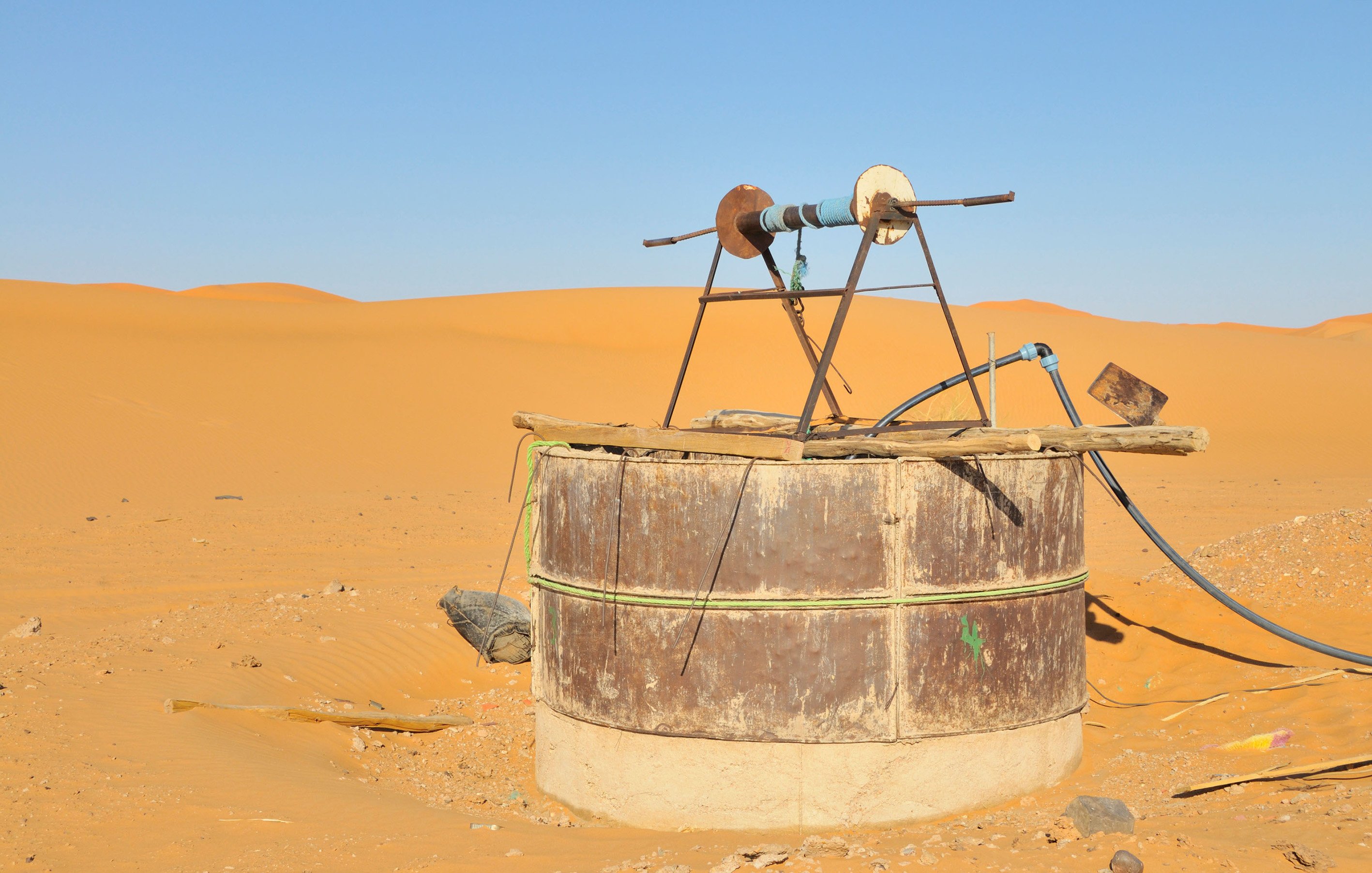 The image shows an old water well situated in a vast desert landscape. The well is made from a large, cylindrical structure with a rusty metal exterior and a concrete base. At the top, there is a simple wooden frame holding a pulley and a couple of wheels, likely used for drawing water. Surrounding the well are sand dunes, displaying a warm golden color under a clear blue sky, emphasizing the arid environment.