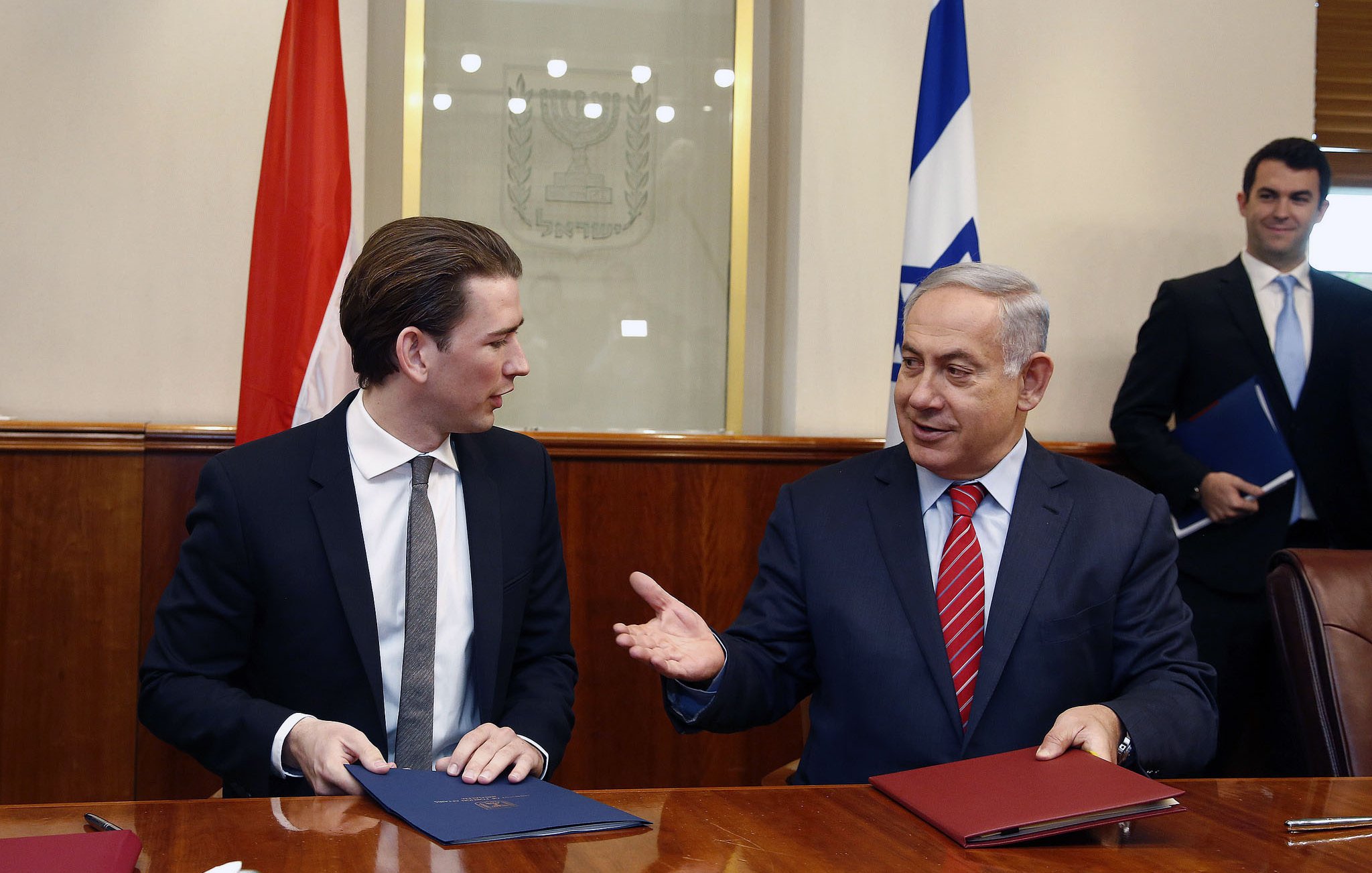 The image shows two men seated at a table in a formal setting. One man, wearing a black suit and tie, appears to be engaged in conversation with the other man, who is wearing a suit with a red tie. They are both holding folders and look to be discussing something important. In the background, there are flags visible, indicating a diplomatic context. The atmosphere appears professional and cooperative.