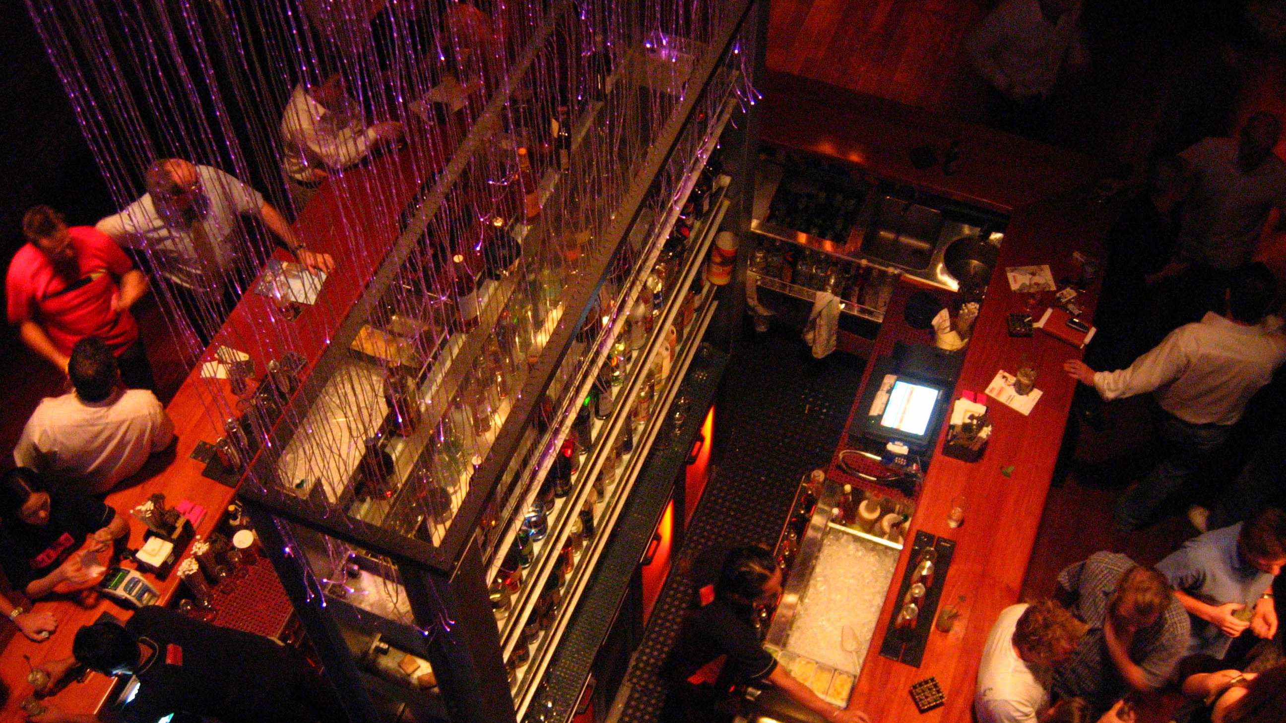 The image depicts a lively bar scene viewed from above. The bar features multiple shelves filled with bottles and is adorned with decorative lights. People are gathered throughout the space, engaging in conversation and enjoying drinks. The atmosphere seems vibrant and social, with a mix of patrons at the bar and tables. The lighting adds a warm, inviting ambiance to the setting.