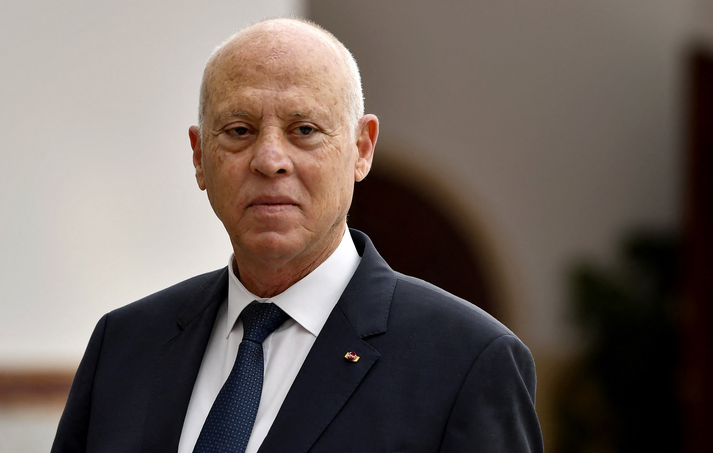 The image shows a man dressed in a suit with a tie, standing in what appears to be an official or dignified environment. He has a serious expression on his face and is looking slightly away from the camera. The background is softly blurred, suggesting it is a formal setting.