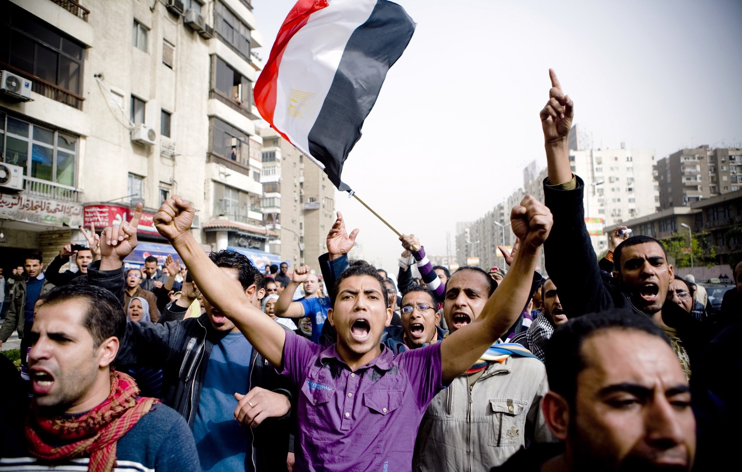 The image depicts a crowd of people in a street, likely engaged in a protest or demonstration. A man in the foreground is holding an Egyptian flag aloft and appears to be shouting passionately. The crowd around him is animated, with several individuals raising their fists and expressing strong emotions. The scene captures a sense of unity and determination among the protestors, amidst urban surroundings with buildings and visible signs of city life.