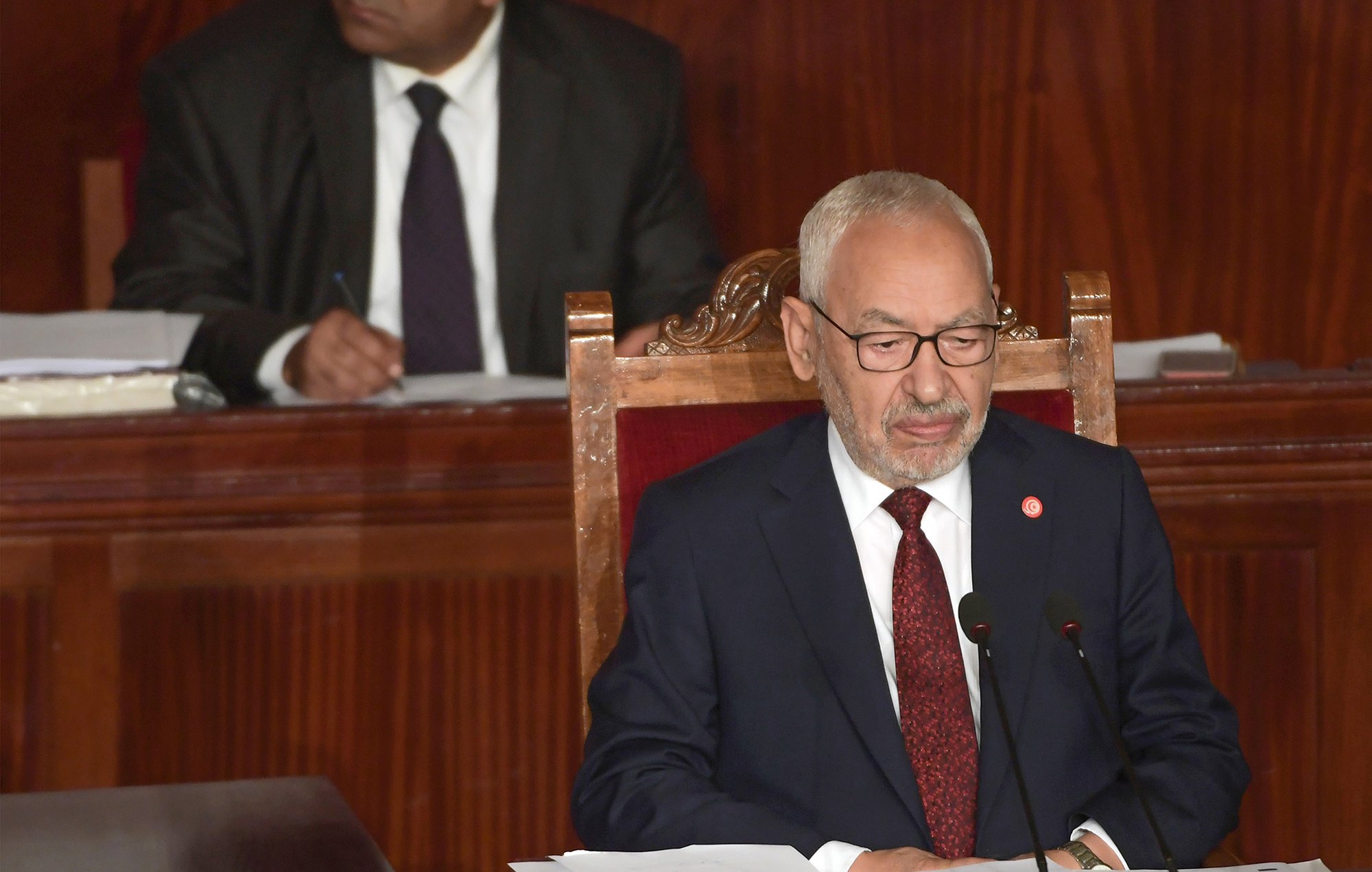 The image shows a man sitting at a podium in a formal setting, likely a legislative or parliamentary assembly. He is wearing a dark suit and glasses, and appears to be speaking or presenting. In the background, another man is seated and observing, dressed in a suit and tie. The environment has a wooden backdrop that contributes to a serious and official atmosphere.