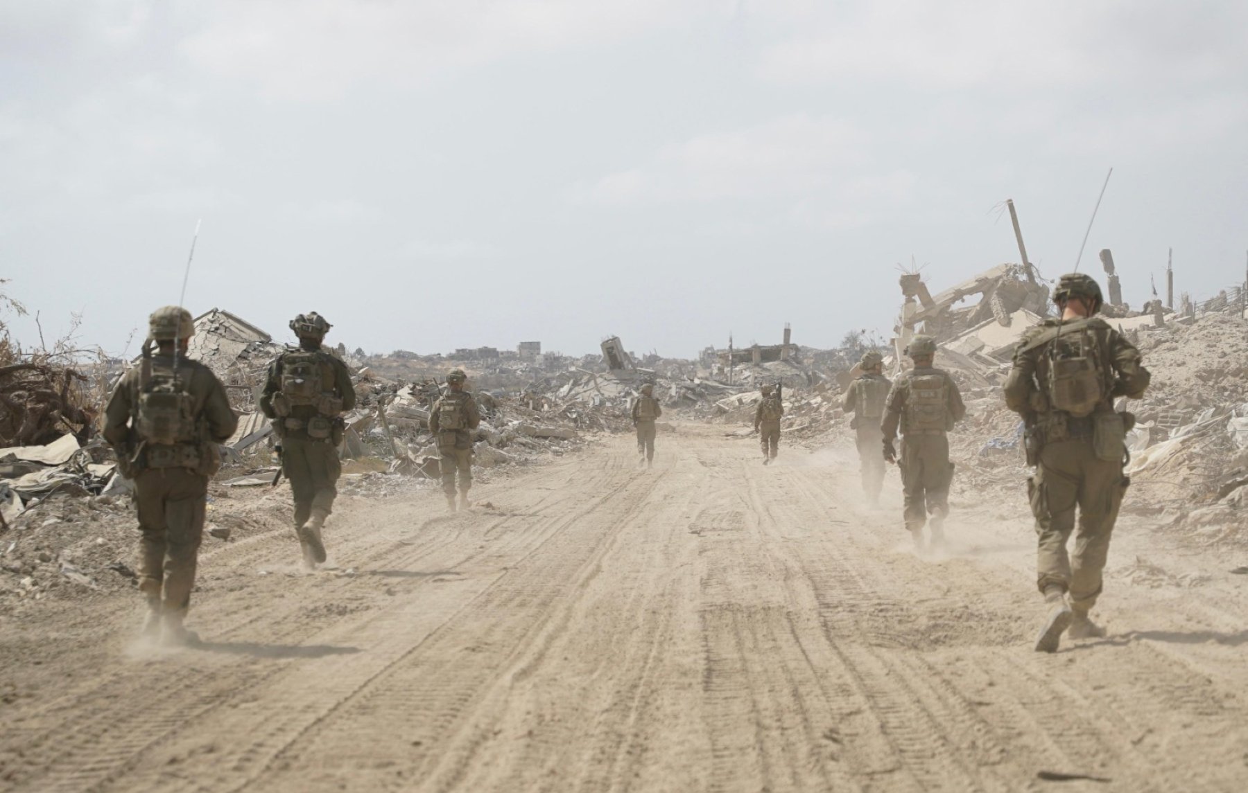 Soldiers in military gear walk through a dusty, desolate landscape with debris around them.