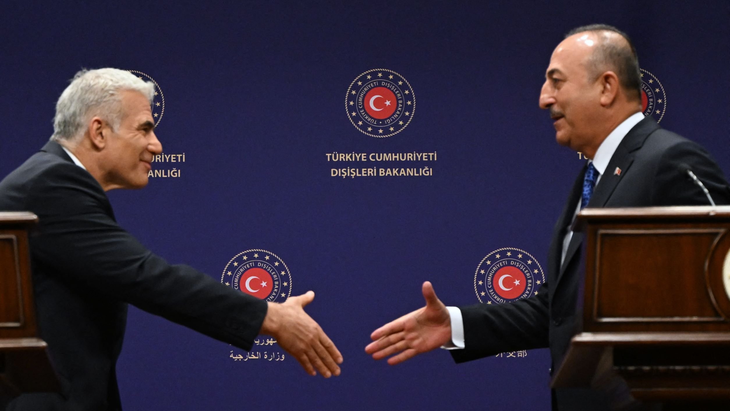 The image depicts two men engaged in a handshake, likely at a diplomatic or formal meeting. They are standing on either side of a wooden podium, with a background featuring the logo of the Turkish Ministry of Foreign Affairs. One man appears to be offering his hand in a gesture of greeting, while the other is responding. Both are dressed in formal attire, suggesting a professional context.