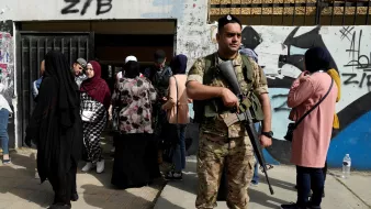 The image shows a soldier wearing military gear and holding a weapon, standing guard in front of a building. In the background, a group of people, predominantly women dressed in various styles of clothing and headscarves, are gathered near the entrance of the building. The setting appears to be an urban area, with graffiti visible on the wall behind the soldier. The atmosphere suggests a controlled environment, possibly in the context of an event or security operation.