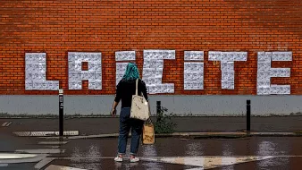 The image shows a person standing in front of a brick wall on which the word "LAICITE" is prominently displayed in large, white, block letters. The individual appears to be wearing a turquoise headscarf and carries a bag, facing the wall. The ground is wet, suggesting it may have recently rained, and the scene includes elements indicating a city environment, such as street signs and a road marking in the foreground. The overall atmosphere combines urban art with a mood of contemplation or engagement with the word on the wall.