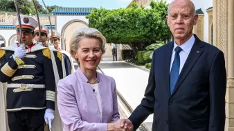 The image shows a formal greeting between two individuals, with one wearing a light purple suit and the other in a dark suit with a tie. They are shaking hands in an outdoor setting, surrounded by greenery and architectural features. In the background, there are guards in ceremonial uniforms standing at attention. The atmosphere appears official and welcoming, suggesting a diplomatic meeting.