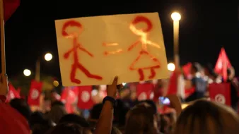 The image shows a night scene of a demonstration or rally, where a person is holding up a sign. The sign features a simplistic representation of a male figure and a female figure, with an equals sign between them, suggesting a message of gender equality. In the background, a crowd can be seen, along with red flags that likely represent a political or social movement. The atmosphere appears to be vibrant and engaged, reflecting a collective message or cause.