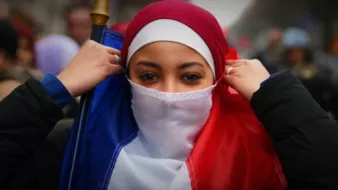The image features a young woman dressed in a red and white outfit, with a hijab, who is adjusting her clothing. She is holding a flag that represents France, with the colors of blue, white, and red. The background suggests a public gathering or demonstration, with people present in a city setting. The atmosphere appears to convey themes of identity and cultural expression.