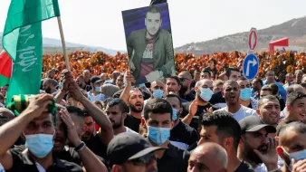 The image depicts a large crowd participating in a protest or demonstration. Many individuals are holding up flags, including green ones, and a prominent figure is holding a portrait of a man. The crowd appears to be engaged and unified, with some people wearing face masks. The background features hills, adding to the overall atmosphere of the event.