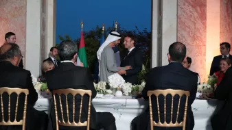 The image depicts a formal gathering, likely a diplomatic event or dinner. In the foreground, several individuals are seated at a long table, facing away from the camera. They appear to be engaged in conversation or listening attentively. In the background, two men are shaking hands or exchanging greetings, with flags visible nearby, suggesting a moment of diplomacy or cooperation. The setting features elegant decor, with floral arrangements on the table and a softly lit ambiance, indicating an evening event.