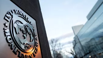 The image features the emblem of the International Monetary Fund (IMF) prominently displayed on a stone surface. The emblem includes a globe surrounded by olive branches, symbolizing peace and cooperation. In the background, there is a modern building with glass windows, reflecting a serene sky with some clouds. The setting appears to be urban and professional, likely outside the IMF headquarters.