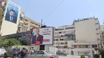 The image shows a street scene featuring a large billboard on a building. The billboard displays a portrait of a man and includes text in Arabic. In the background, there are other buildings, some of which also feature large advertisements. A couple of vehicles, including a motorcycle and a car, are visible on the road. The sky is clear and blue, indicating a sunny day.
