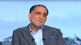 A middle-aged man in a suit with a serious expression, seated against a blurred background.