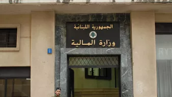 The image shows a soldier standing outside a building with a large sign displaying the words "الجمهورية اللبنانية" (Lebanese Republic) and "وزارة المالية" (Ministry of Finance) in Arabic. The building has a modern façade with a marble entrance. The soldier is wearing military uniform and holding a weapon, indicating a security presence at the site. The overall setting suggests a government location in Lebanon.