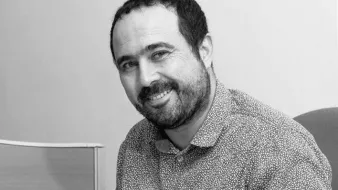 The image shows a man sitting and smiling. He has a short beard and is wearing a spotted button-up shirt. The background appears to be a plain wall and a piece of furniture, suggesting a professional or office setting. The overall tone of the image is friendly and approachable.