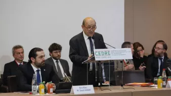 The image depicts a formal conference setting. A middle-aged man, presumably a speaker, is standing at a podium with a sign that reads "CEDRE" and indicates the location and date of the event. He appears to be addressing an audience that includes several men seated behind him, some of whom are engaged in listening or taking notes. The environment has a professional tone, with tables set up and various drinks and papers visible. The atmosphere suggests a significant discussion or presentation, likely focused on economic or policy-related matters.