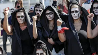 The image depicts a group of people wearing black hooded cloaks and painted faces, featuring white and dark makeup that gives a striking visual effect. They appear to be raising their fists in a gesture of solidarity or protest. The atmosphere suggests they are part of a demonstration or performance, possibly conveying a message or advocating for a specific cause. The backdrop includes hints of a larger gathering, emphasizing the collective nature of their presence.