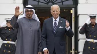 The image shows two men standing at the entrance of a building, likely the White House. One man is dressed in traditional attire with a white headscarf, while the other is in a formal suit. They are both smiling and posing for the camera, with one man waving and the other giving a thumbs-up. In the background, there are uniformed guards standing at attention. The scene conveys a sense of diplomacy and cordiality between the two leaders.